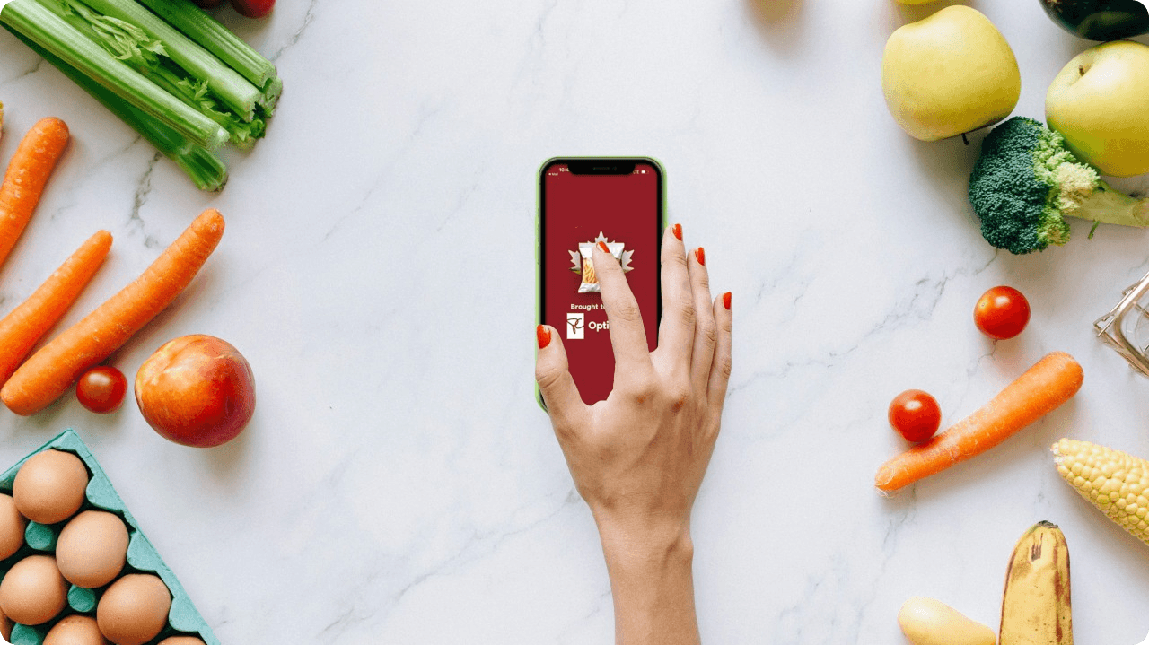 Hand holding a smartphone displaying a sponsored PC Optimum app takeover, surrounded by fresh groceries including carrots, celery, apples, broccoli, eggs, and tomatoes, illustrating a retail media campaign within a grocery shopping context.